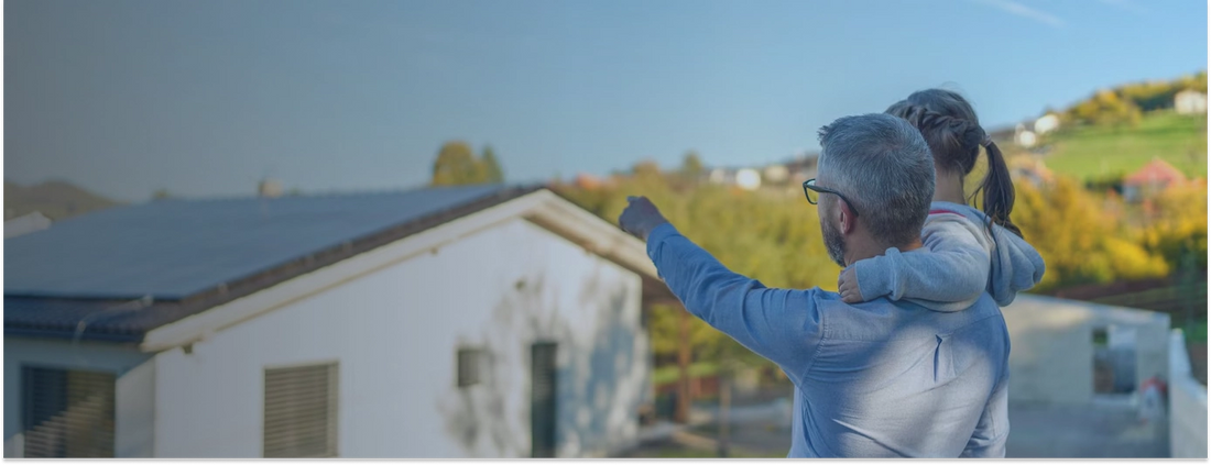 A man with gray hair and glasses holds a young child while pointing toward a white house with solar panels on the roof, with a green landscape and trees in the background.