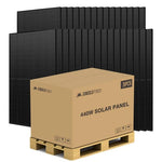 A cardboard box labeled 440W Solar Panel sits on a wooden pallet in front of multiple black solar panels standing upright in the background, its bold ink lettering clearly visible against the box’s surface.