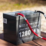 A gray Rich Solar LiFePO4 Battery, 12.8V 100Ah (1280Wh), with red and black cables on its terminals, is placed outdoors on a brown surface.