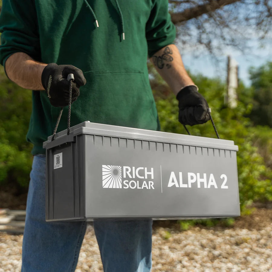 Wearing gloves and a green hoodie, a person carries a large gray Rich Solar ALPHA 2 LiFePO4 Battery (200Ah, 2560Wh) outdoors, with greenery and gravel in the background.