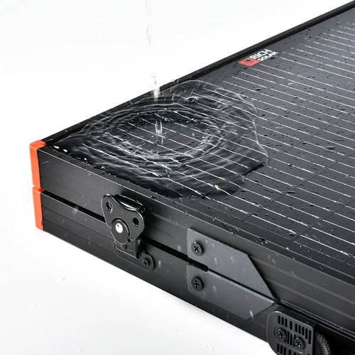 A stream of water splashes onto a closed, black, hard-shell Rich Solar portable case with orange corners and metal latches—built to protect your high-performance solar panel on any off-grid adventure.