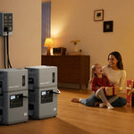 A woman and a girl sit by a decorated Christmas tree surrounded by gifts. Nearby, two large backup power units—including the Mango Power E 3,000W 7kWh 120V Home Backup Portable Power Station from Mango Power—are plugged into a wall outlet. The room is warm and softly lit, creating a cozy atmosphere.