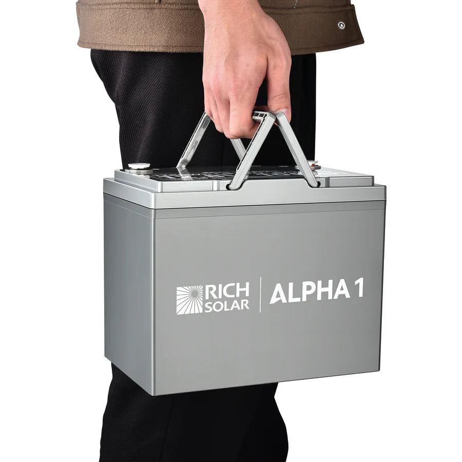 A person holding a gray Rich Solar ALPHA 1 100Ah LiFePO₄ Battery by its handles, standing against a white background.